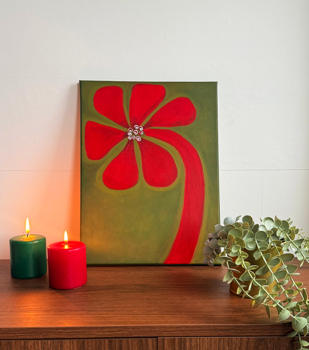 Red flower painting displayed on a wooden sideboard beside red and green candles and a trailing plant.