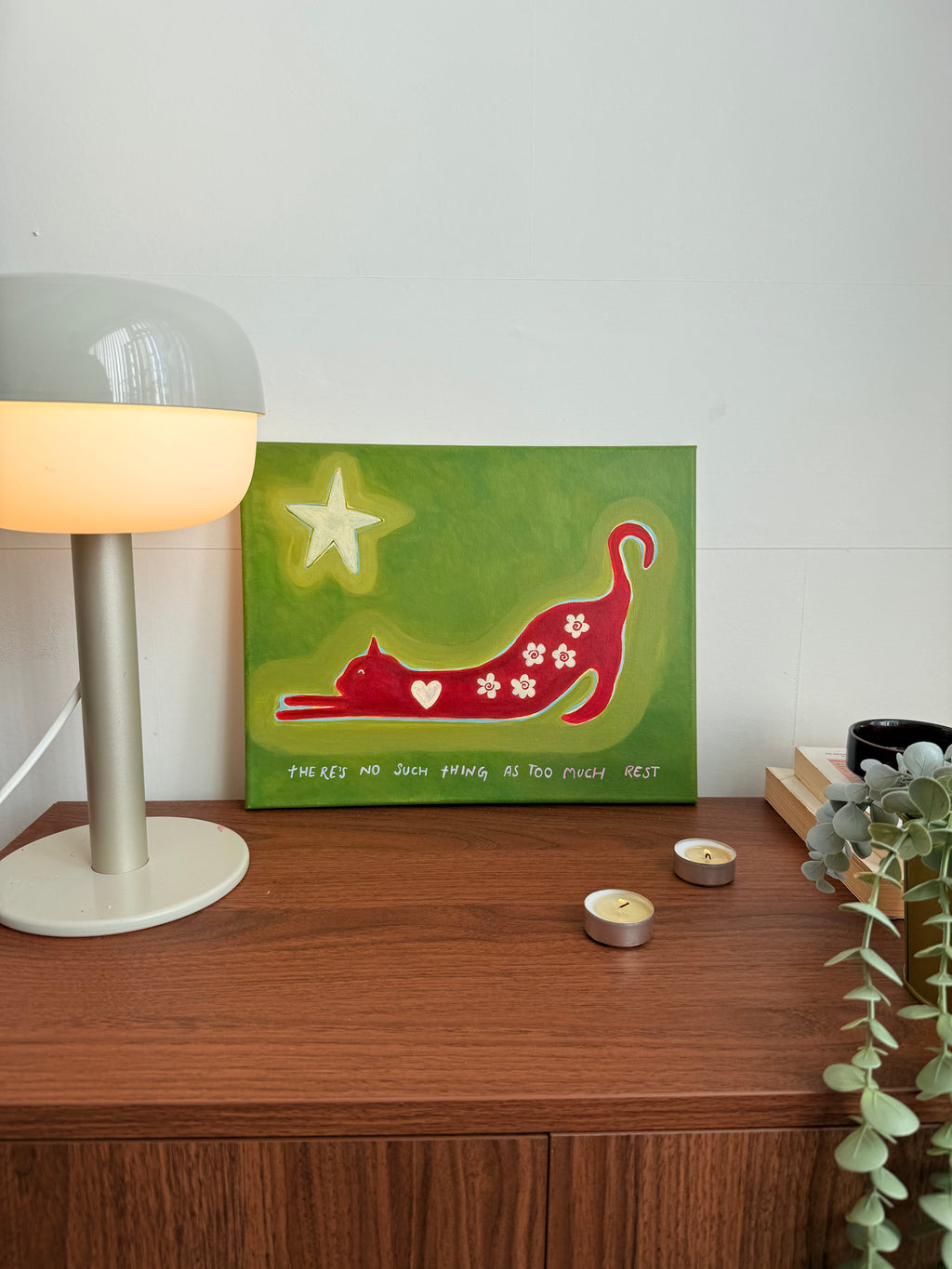 Original red cat artwork on green canvas styled on a wooden cabinet with a trailing plant