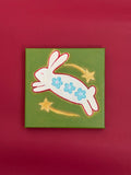 Mini original painting of a leaping white bunny decorated with three blue flowers, surrounded by two golden shooting stars on olive green.
