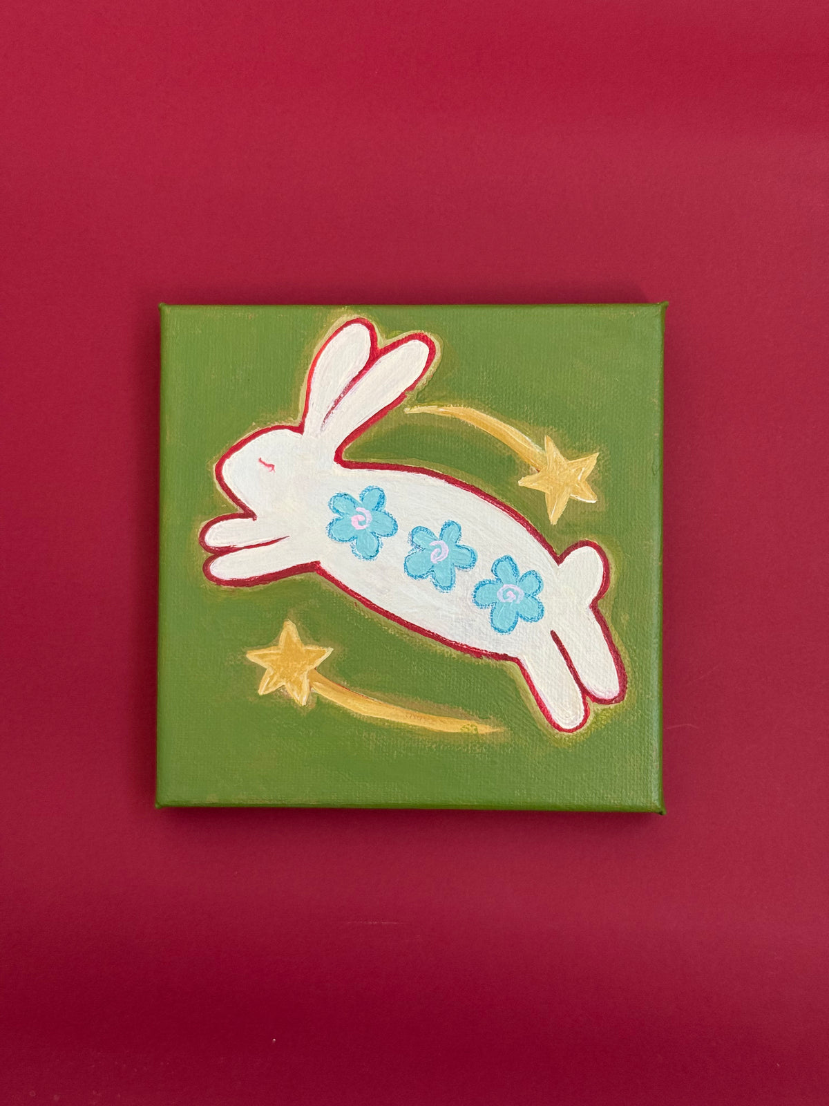 Mini original painting of a leaping white bunny decorated with three blue flowers, surrounded by two golden shooting stars on olive green.