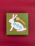 Original painting of a seated white bunny decorated with three blue flowers beneath a golden shooting star on an olive-green background.