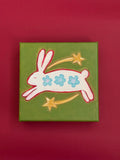 Original painting of a white bunny with three blue flowers, leaping between two golden shooting stars on an olive-green background.