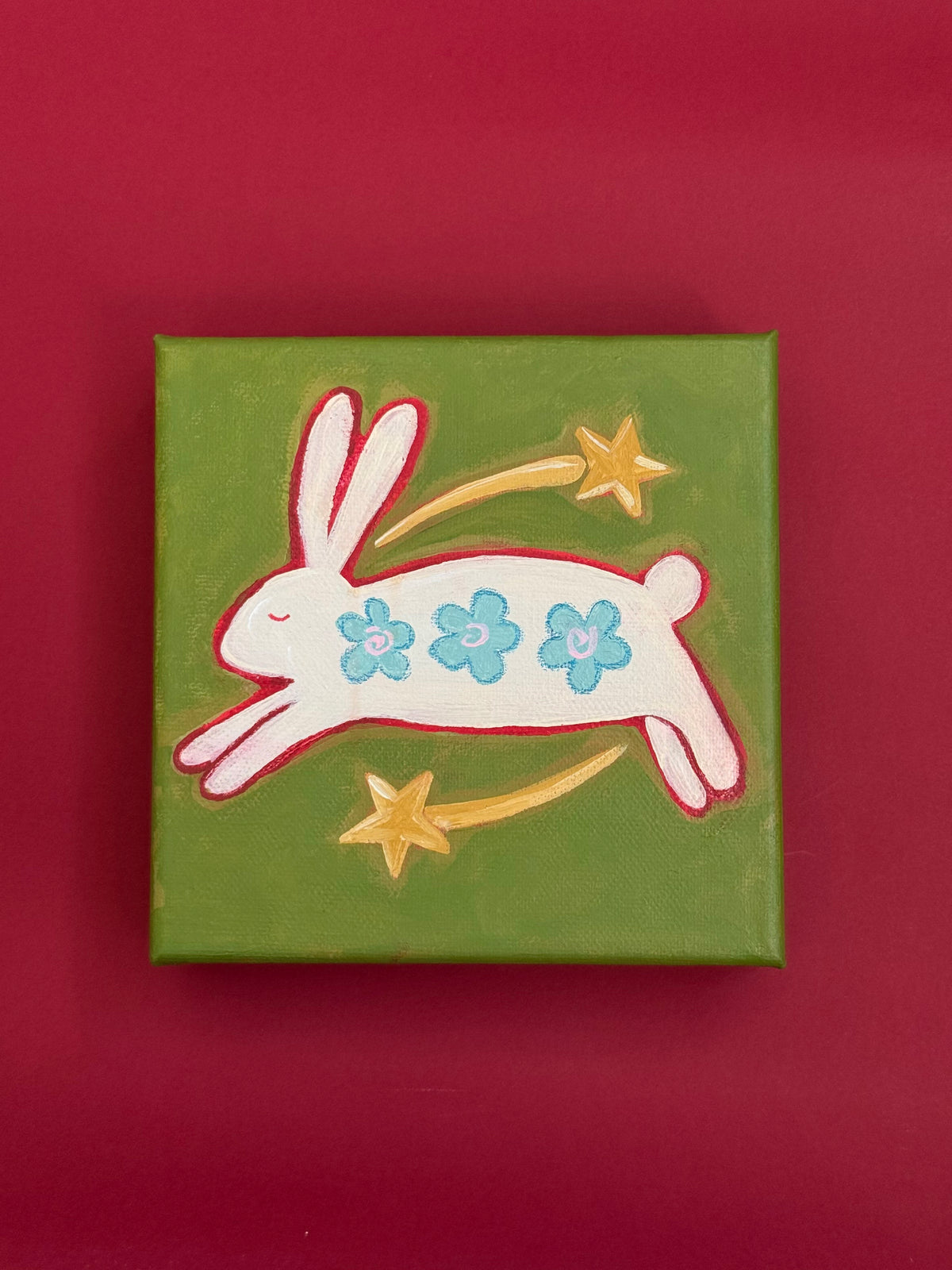Original painting of a white bunny with three blue flowers, leaping between two golden shooting stars on an olive-green background.